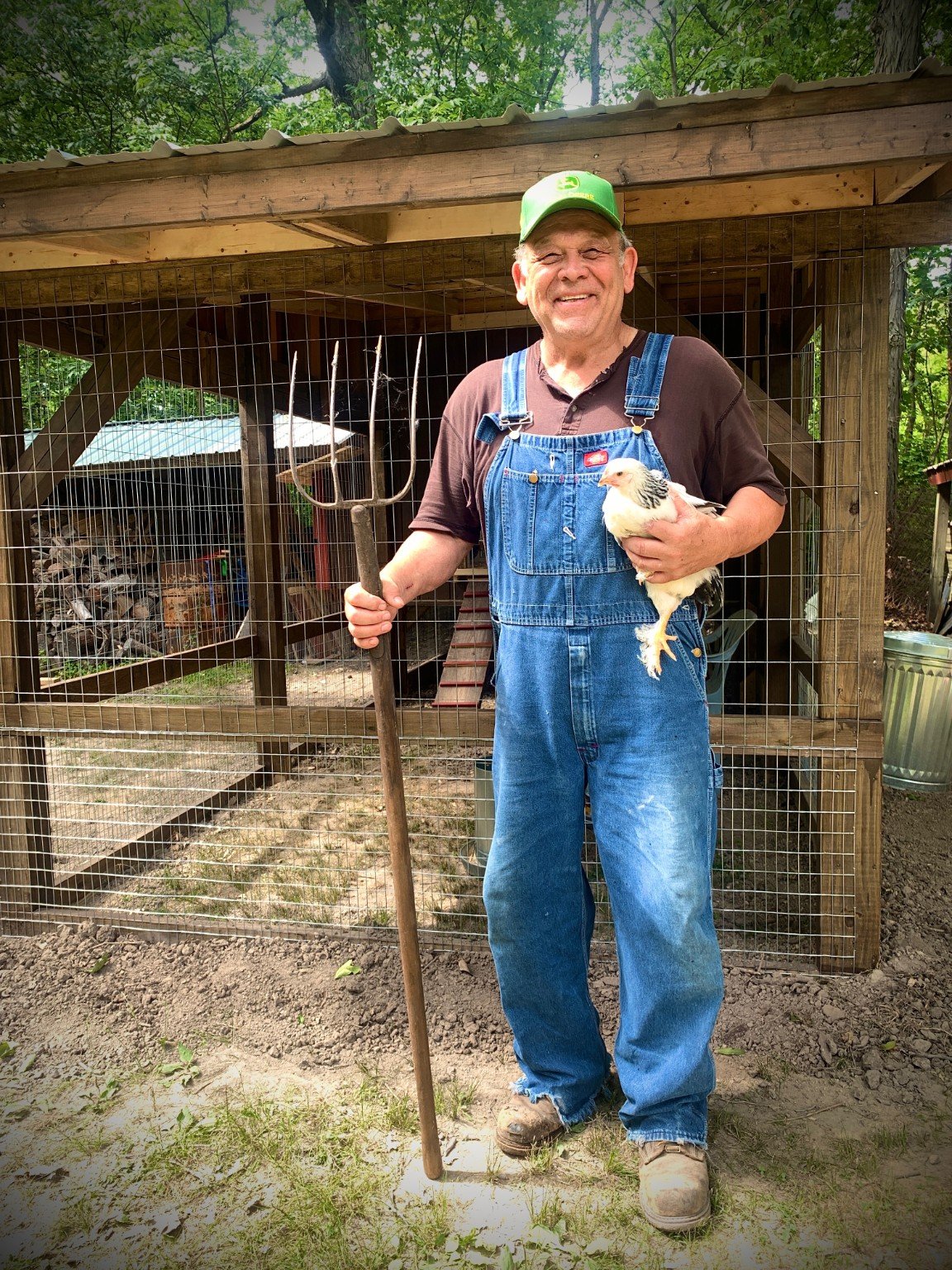 Farmer in overalls holding a chicken beside a handmade coop